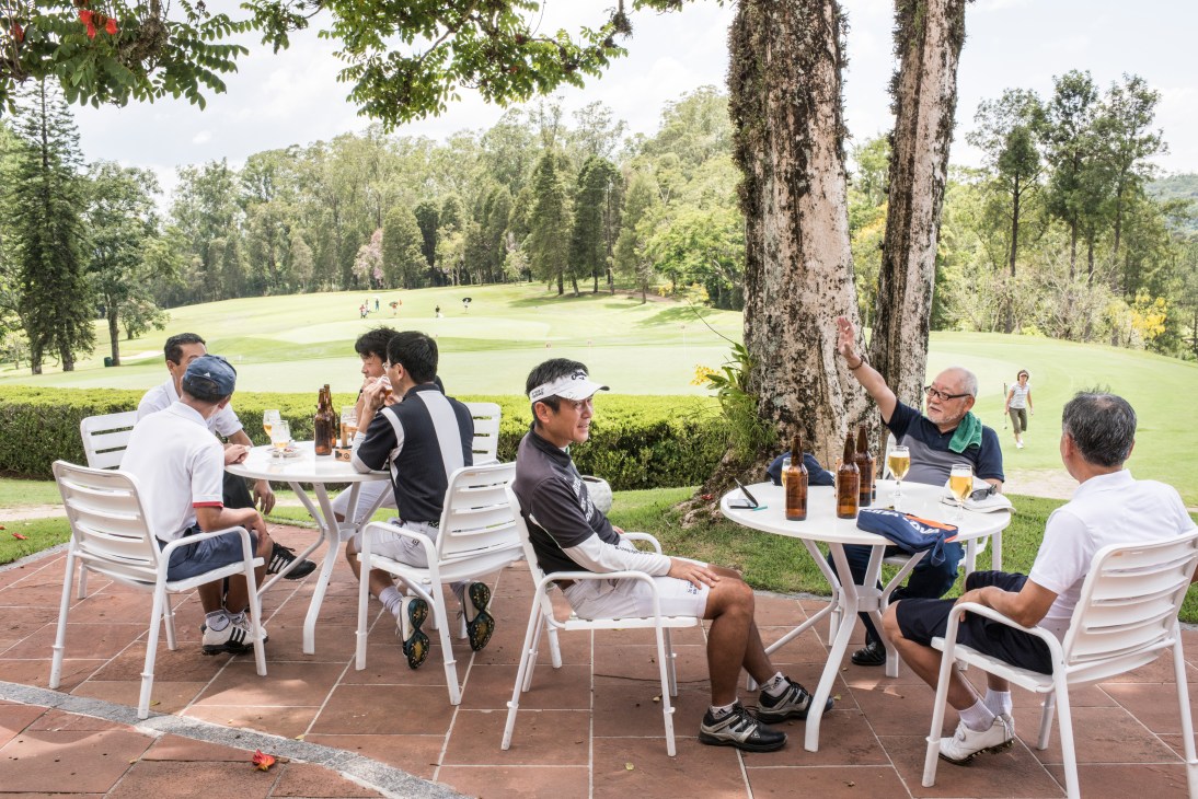 People socializing with drinks at outdoor tables overlooking a golf course with tall trees.