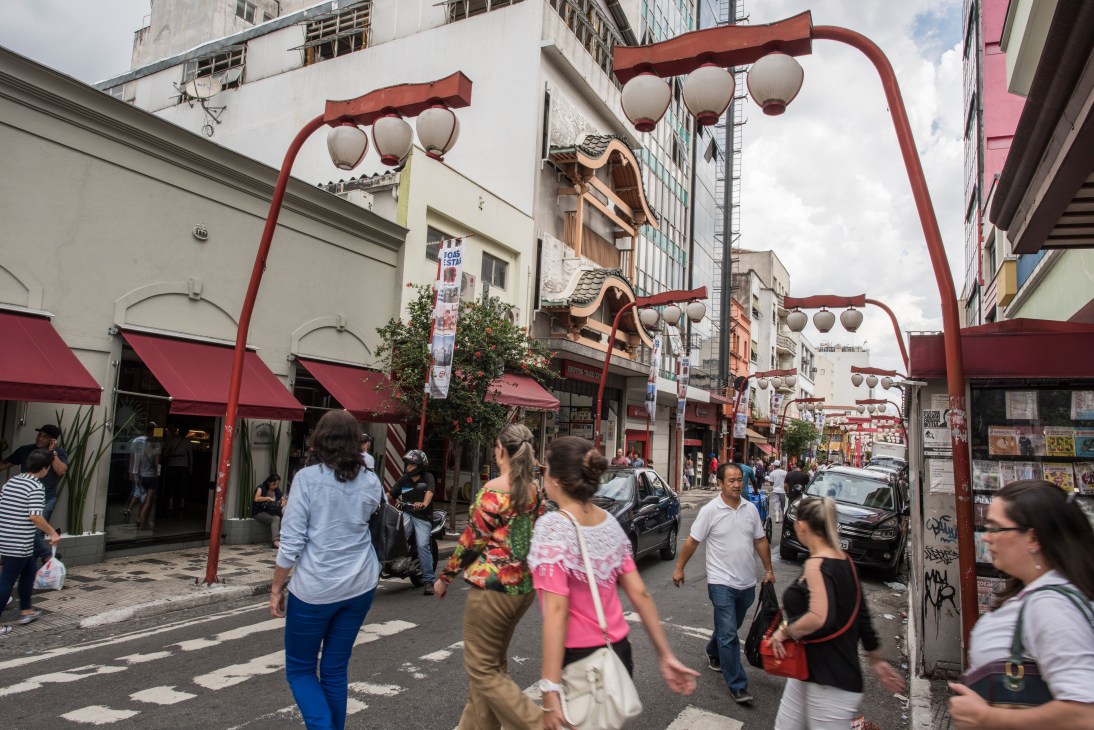 Pedestrians walking through Liberdade neighbourhood's street lined with red lanterns and traditional Japanese architecture in São Paulo.