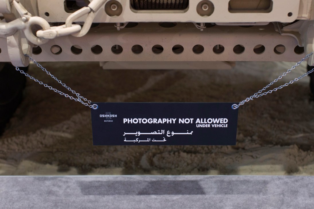 A sign hanging from an Oshkosh Defense military vehicle reading "Photography Not Allowed Under Vehicle" in English and Arabic.