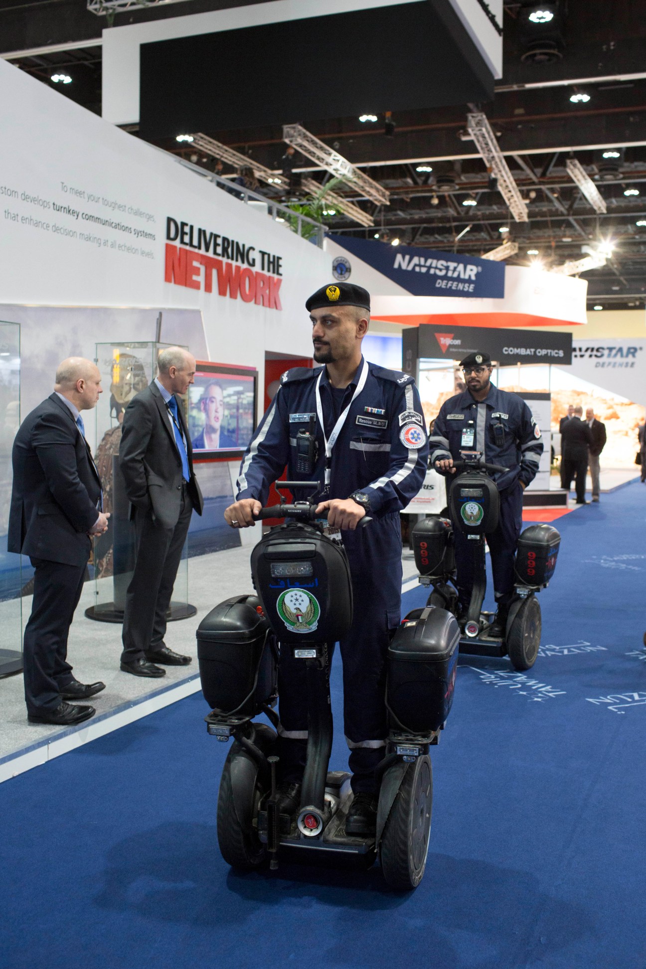 Military personnel on Segways at an indoor defense exhibition trade show.