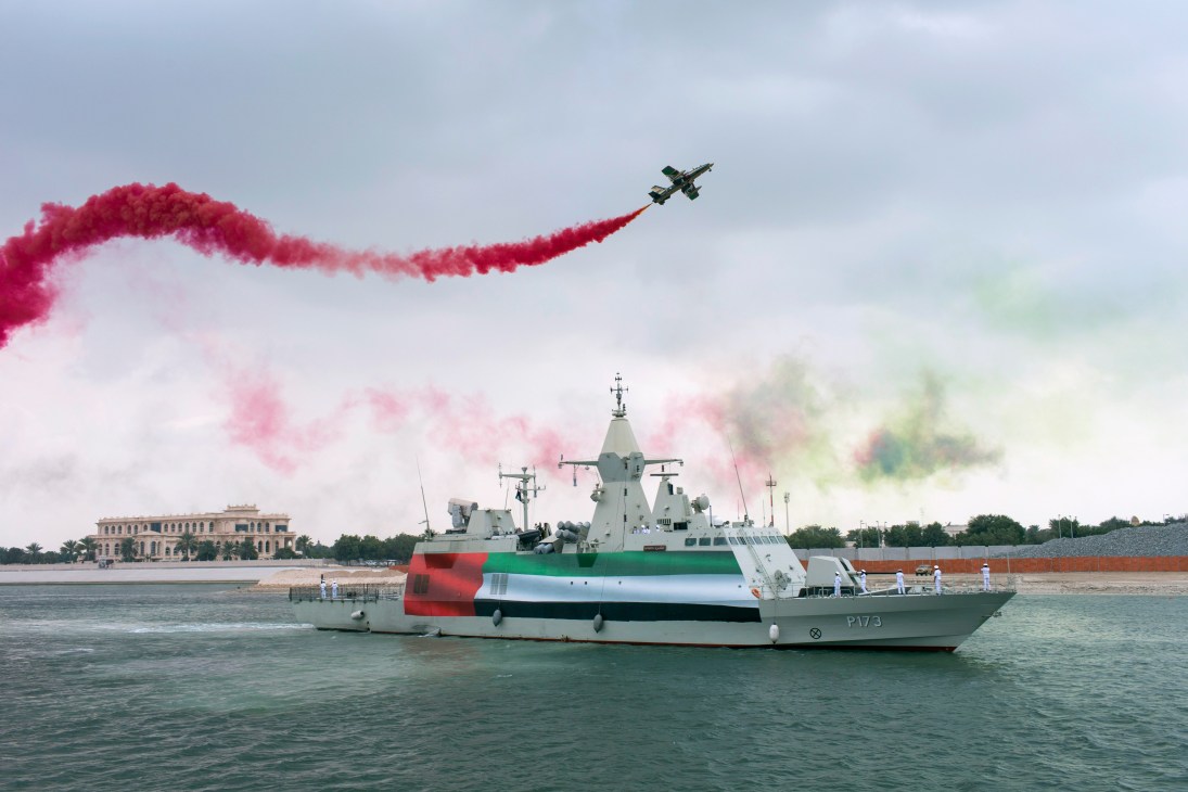 Military jet aircraft performing aerobatics with red smoke trail over UAE naval vessel in water.