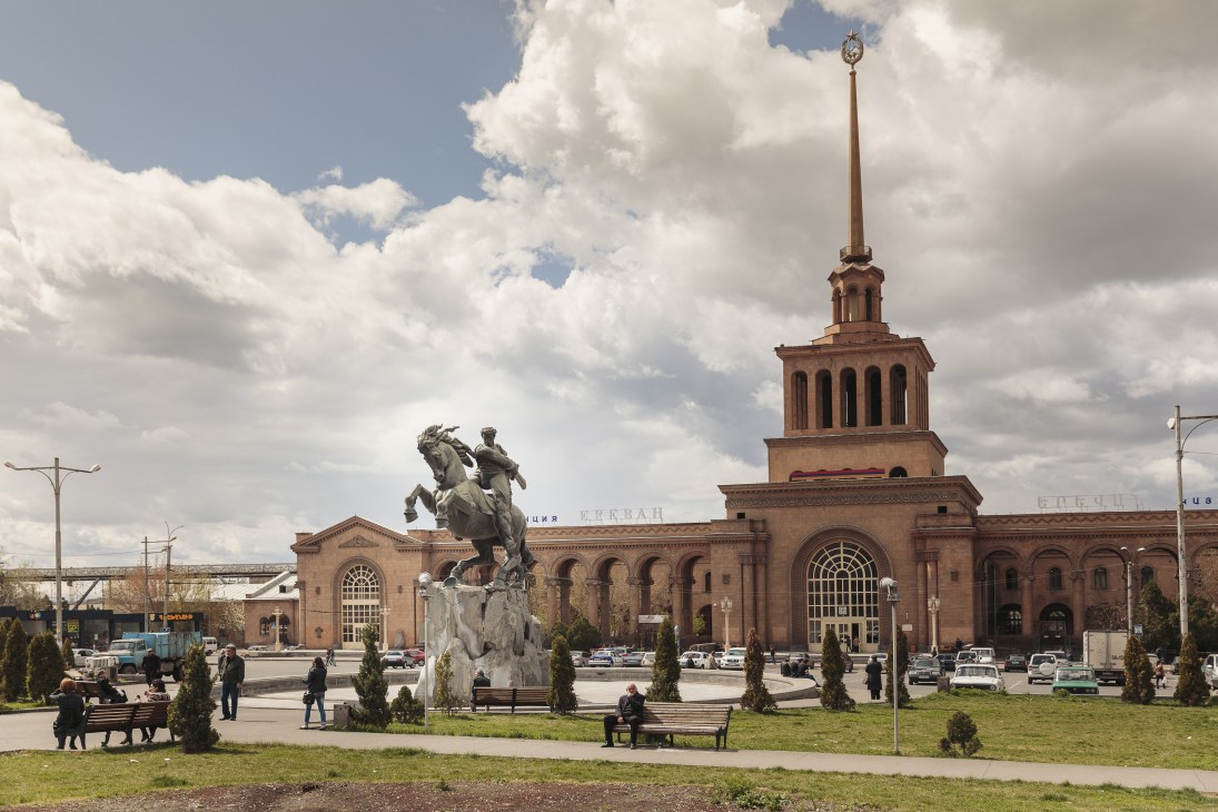 Yerevan railway station with equestrian statue and rose-colored tuff building under cloudy sky.