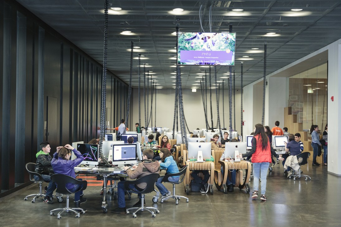 Young students work at Apple Mac computers in a modern, open-plan technology center with industrial ceiling design.