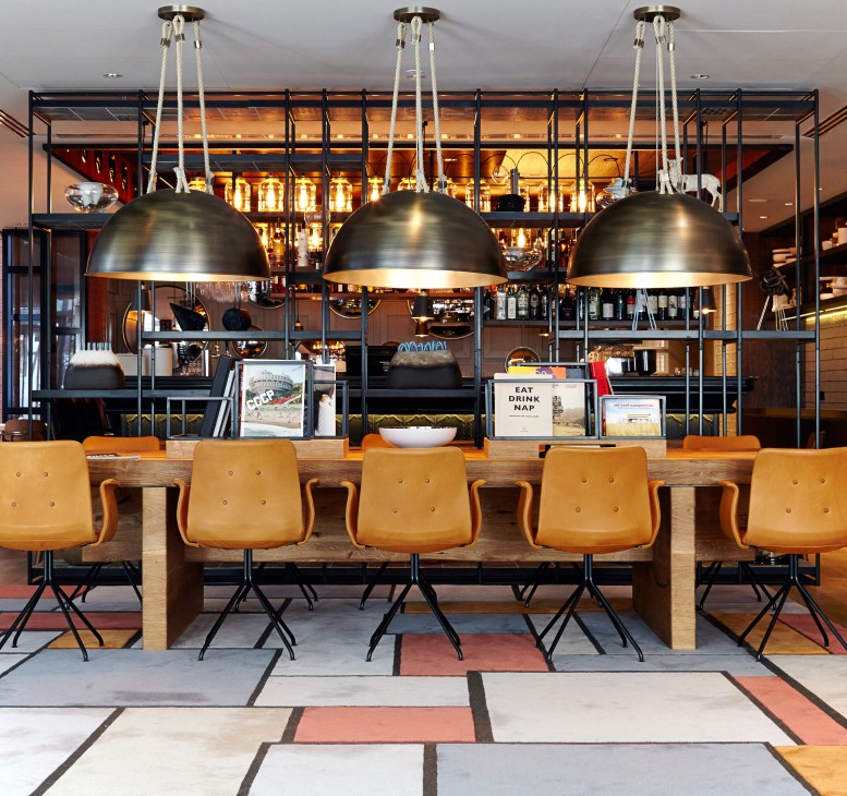 Modern hotel lobby with wooden counter, mustard-yellow chairs, and three large metallic pendant lights.