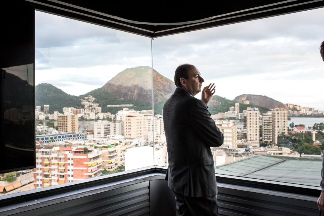 Man in dark suit stands at large window overlooking Rio de Janeiro with mountains and buildings visible behind him.