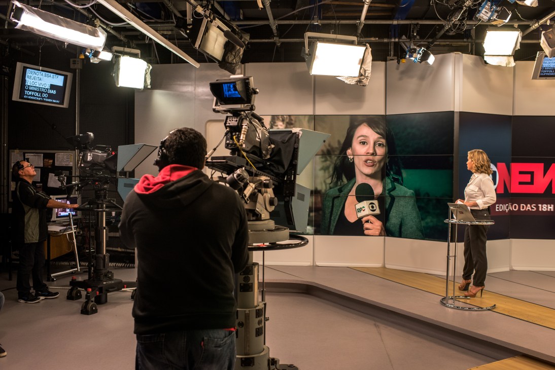 Live broadcast studio at GloboNews with camera operators, presenter at desk, and large monitors displaying the news program.