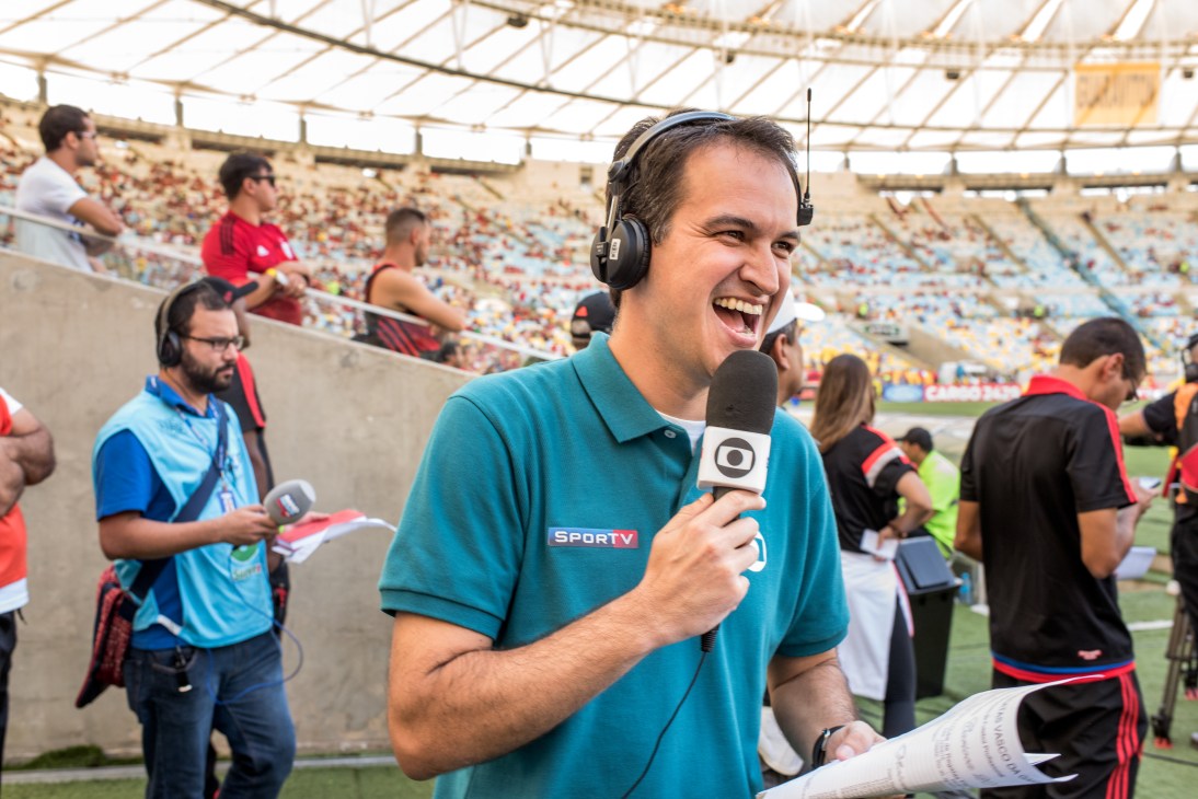 Reporter Edson Viana wearing headphones and SPORTV polo shirt speaks into microphone at Maracanã stadium during live broadcast.