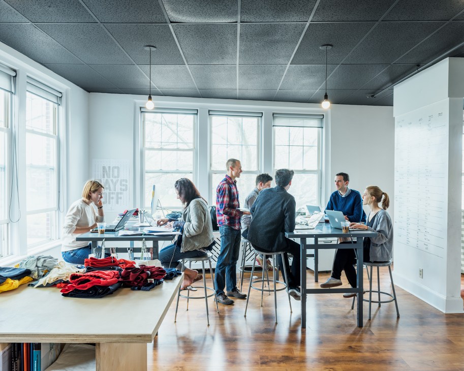 Tracksmith team members work at desks in a bright, modern office with large windows and colorful athletic gear displayed on tables.