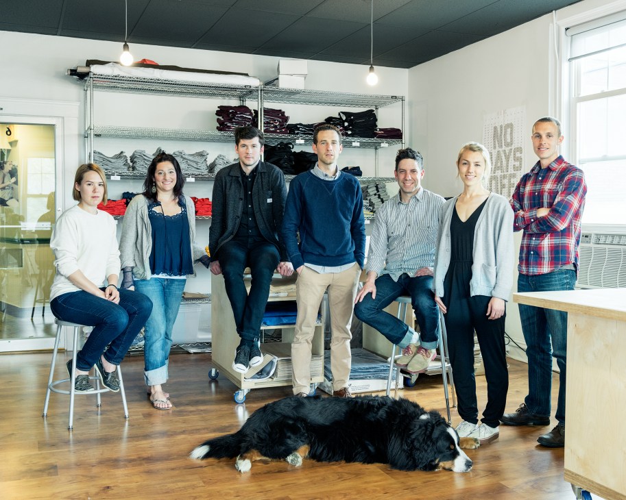Seven Tracksmith team members pose in their Wellesley headquarters with shelving of athletic wear behind them and a black dog lying on the wooden floor.