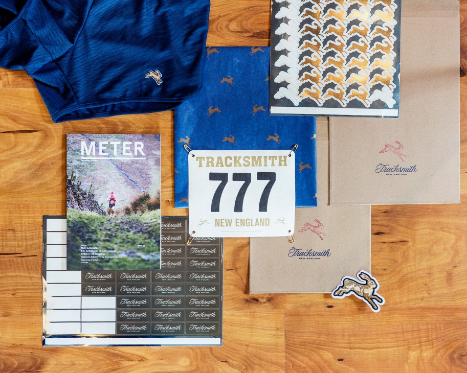 Tracksmith branding materials including Meter Magazine, bib number 777, athletic wear, and design samples arranged on a wooden surface.