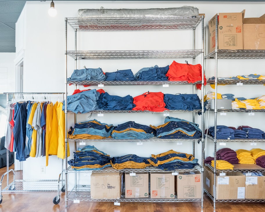 Industrial shelving units displaying neatly organized Tracksmith athletic wear in blue, red, and yellow colors with cardboard boxes below.