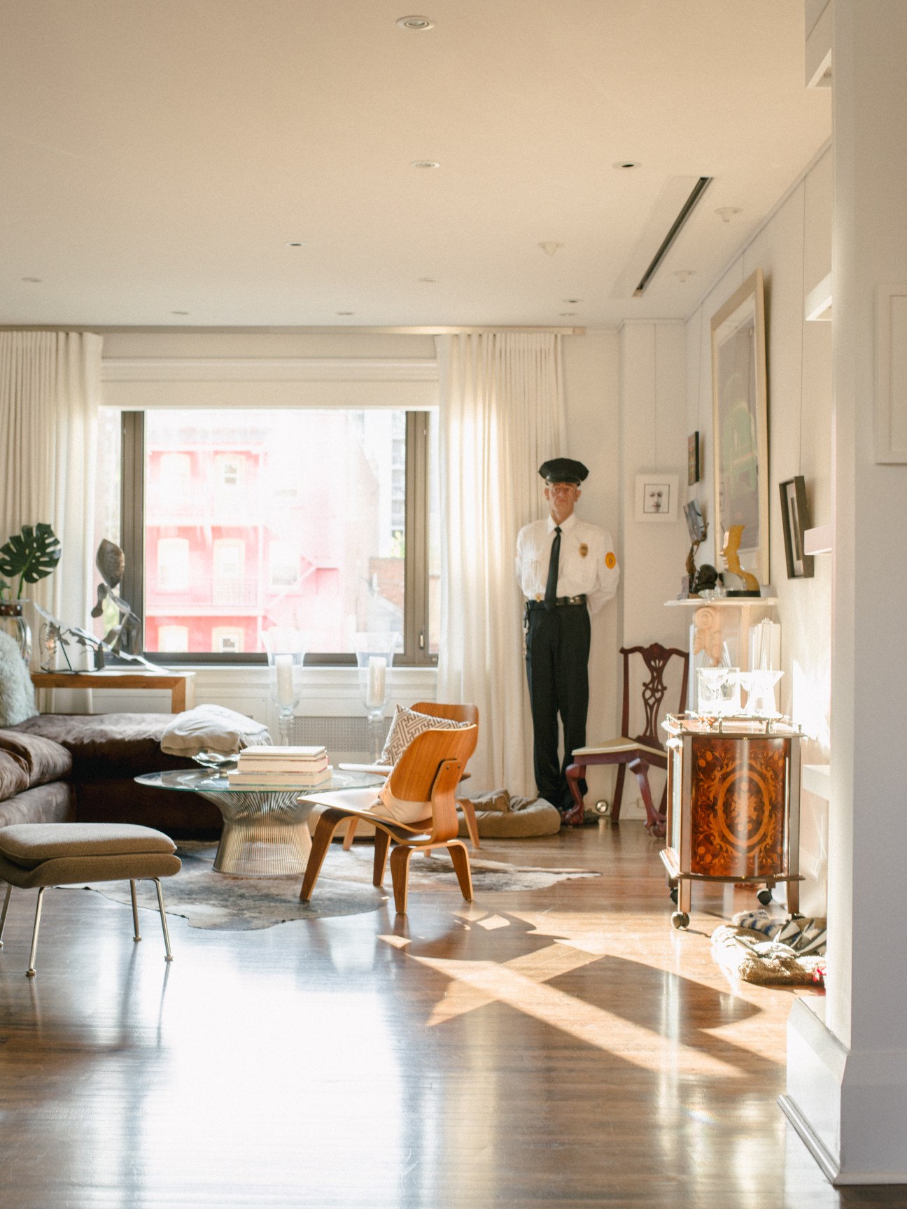 A sunlit modernist living room with wooden floors, mid-century furniture, and a mannequin in formal attire standing by shelving.