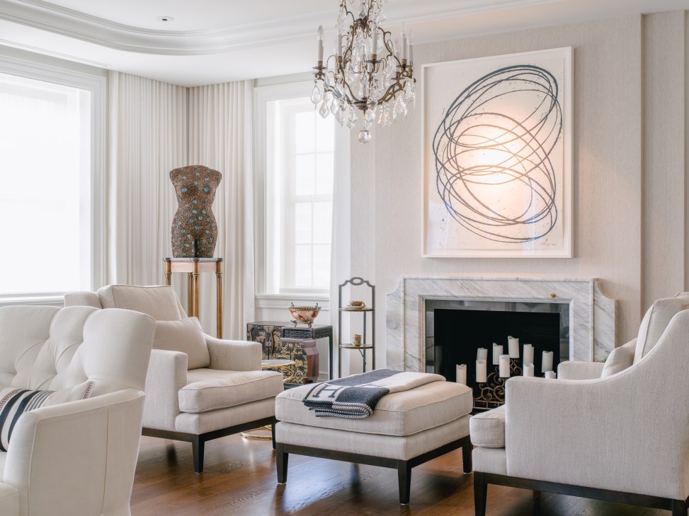 Elegant living room with cream upholstered chairs, fireplace, crystal chandelier, and abstract art above mantelpiece.