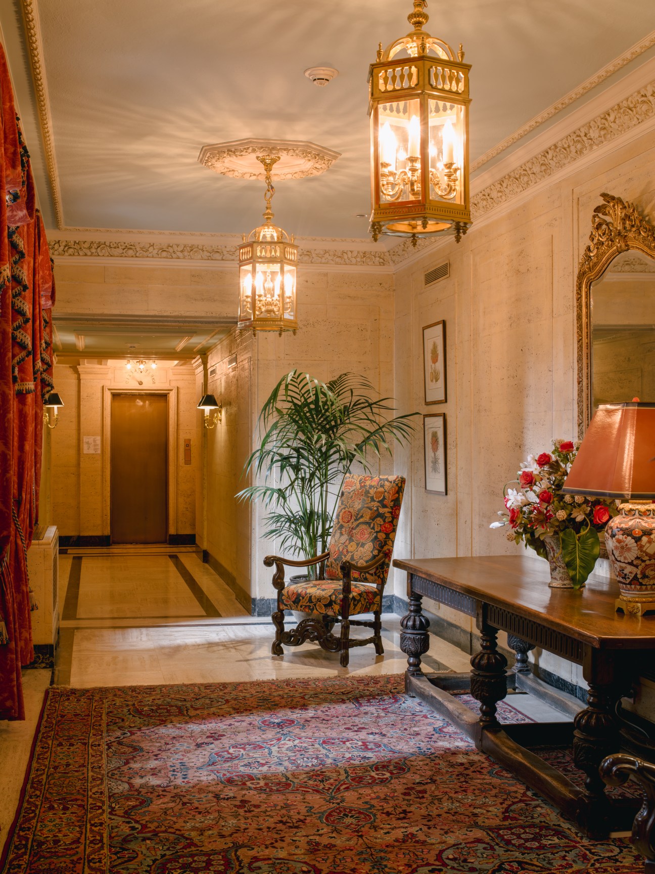 Ornate entrance hall with brass pendant lights, decorative mirror, patterned rug, upholstered chair, and floral arrangement.