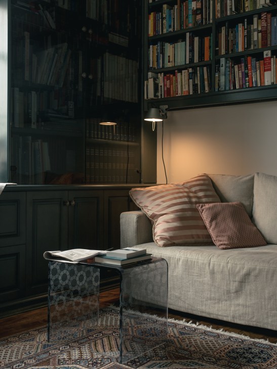 Cozy reading nook with beige sofa, striped and patterned cushions, metal side table with books, and Artemide Tolomeo clamp lamp beside dark built-in bookshelves.