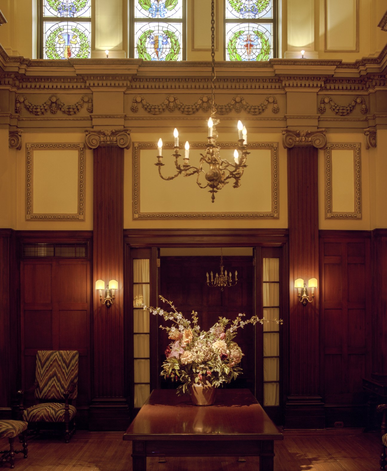 Ornate reception room with stained glass windows, decorative cornicing, brass chandelier, and floral arrangement on table.