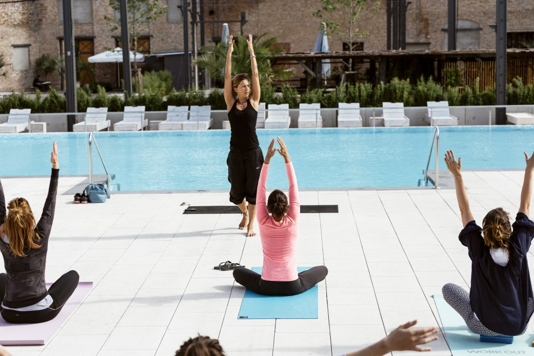 Yoga instructor leading a poolside class at Haubentaucher with participants on mats and white lounge chairs visible behind the pool.