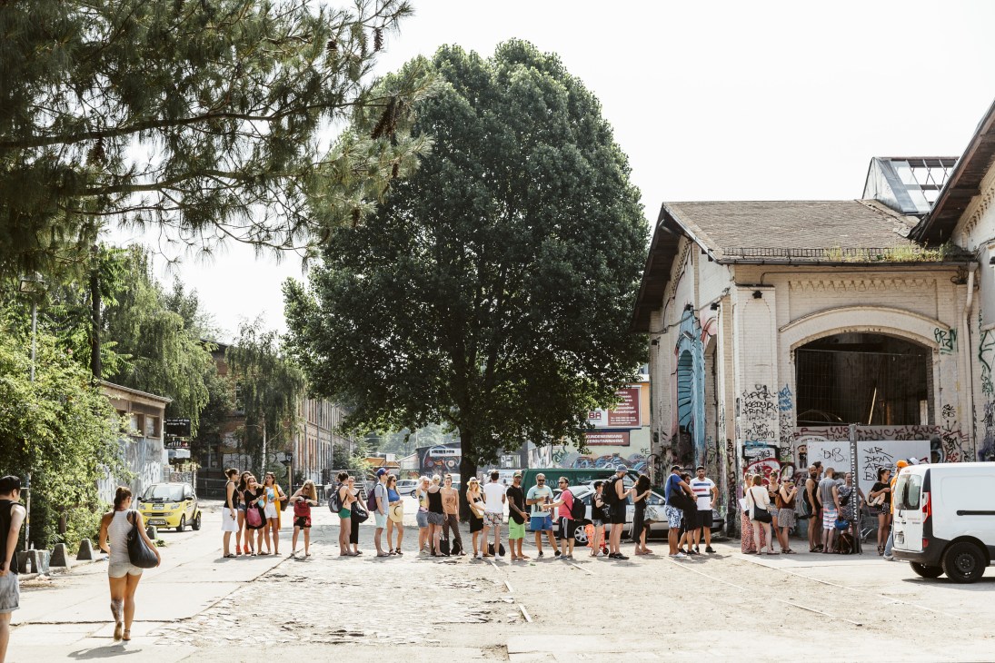 Long queue of people waiting outside industrial brick buildings to enter Haubentaucher pool at Berlin's RAW-Gelände.