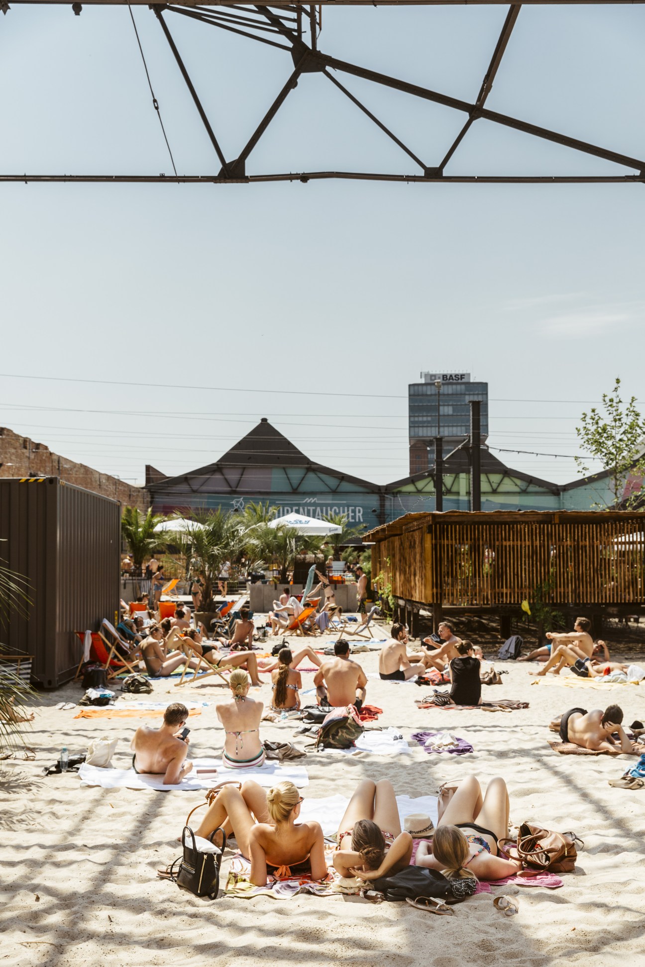 Beachgoers lounging on sand at Haubentaucher's beach area with pool complex and industrial buildings behind.
