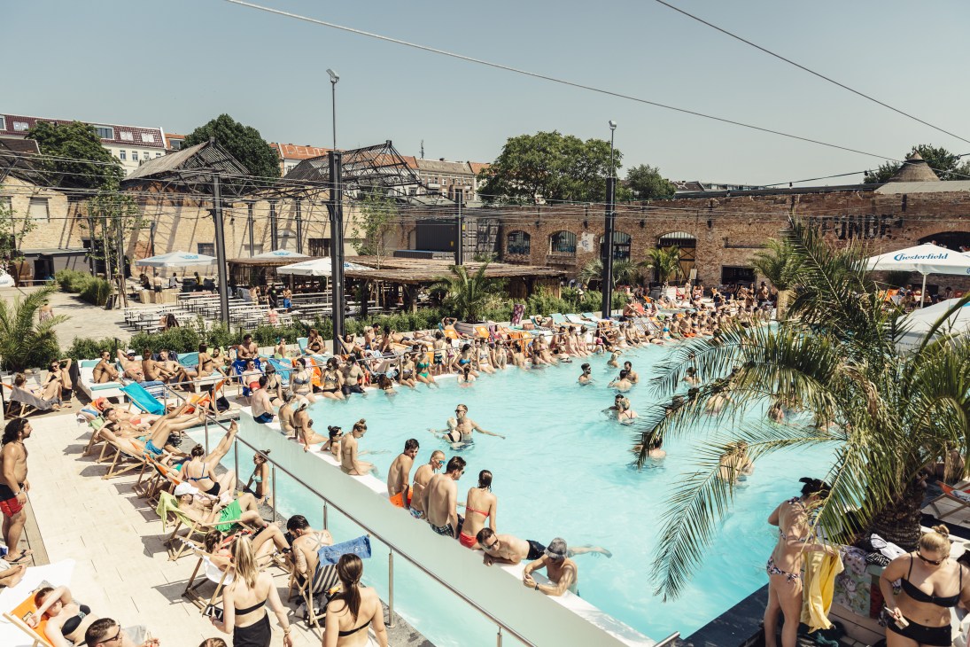 Crowded outdoor swimming pool with sunbathers on lounge chairs and swimmers in turquoise water at Haubentaucher Berlin lido.