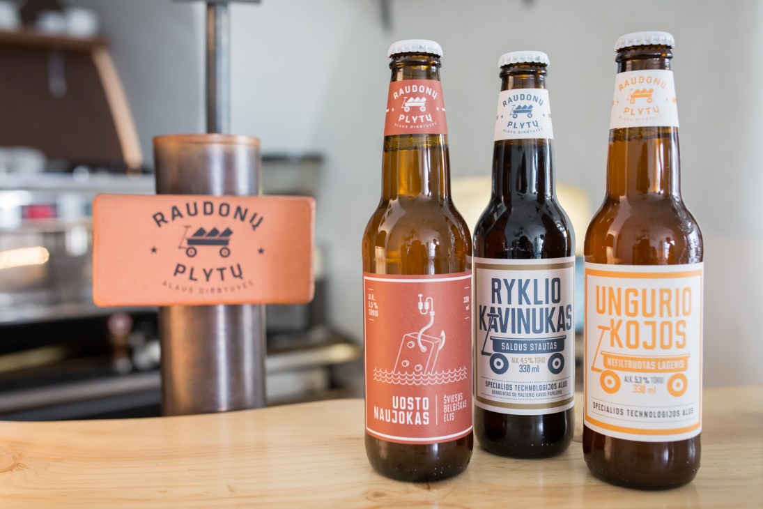 Three bottles of Raudonu Plytu Lithuanian beer with orange and white labels on a wooden counter.