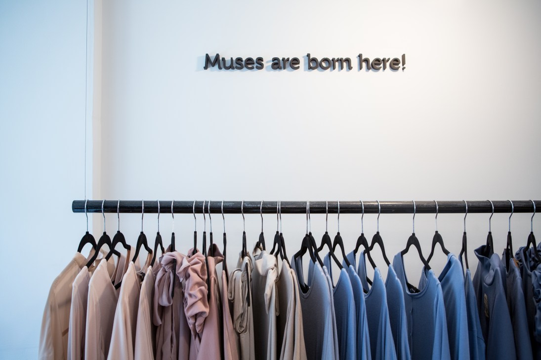 Clothing boutique LeMuse interior with pastel garments on black hangers along a dark rod against white wall with text reading 'Muses are born here!'