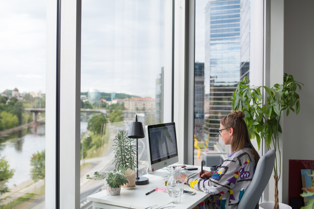 Woman working at computer in bright office with city views and potted plants by floor-to-ceiling windows.