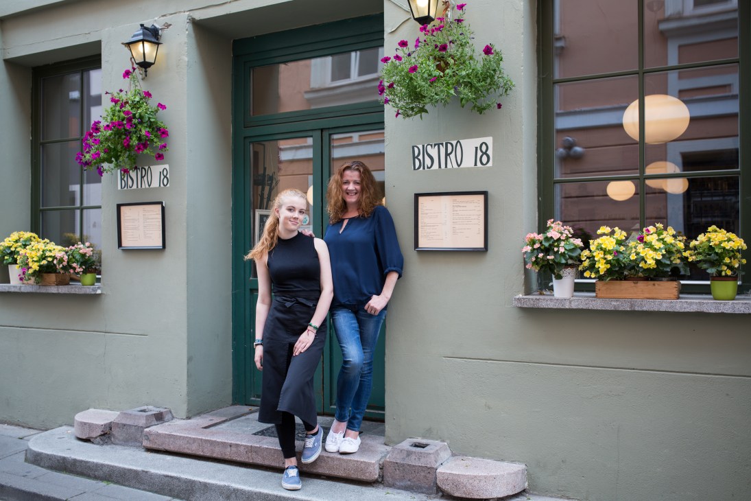 Two women stand outside Bistro 18, a restaurant with green doors and hanging flowering baskets on a white building facade.