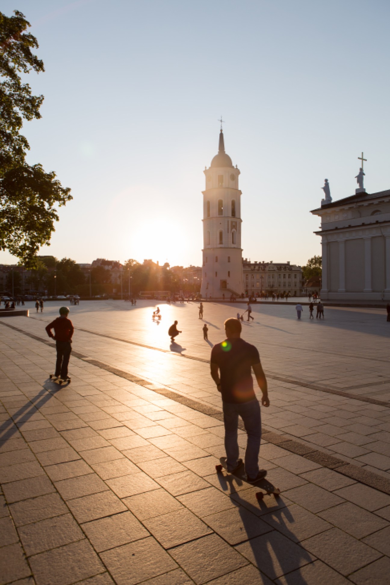 People skateboarding and walking in Cathedral Square at sunset with a pink bell tower and cathedral in the background.