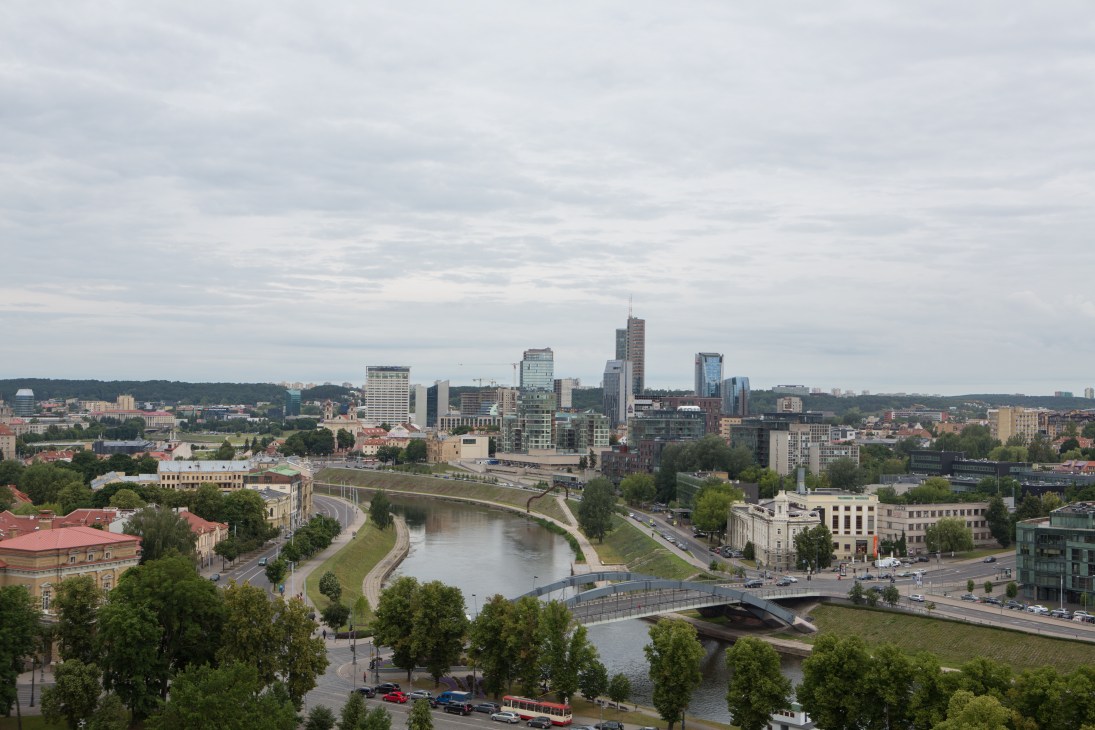 Vilnius cityscape with modern business district skyscrapers, river, bridge, and historic old town with red-tiled roofs.