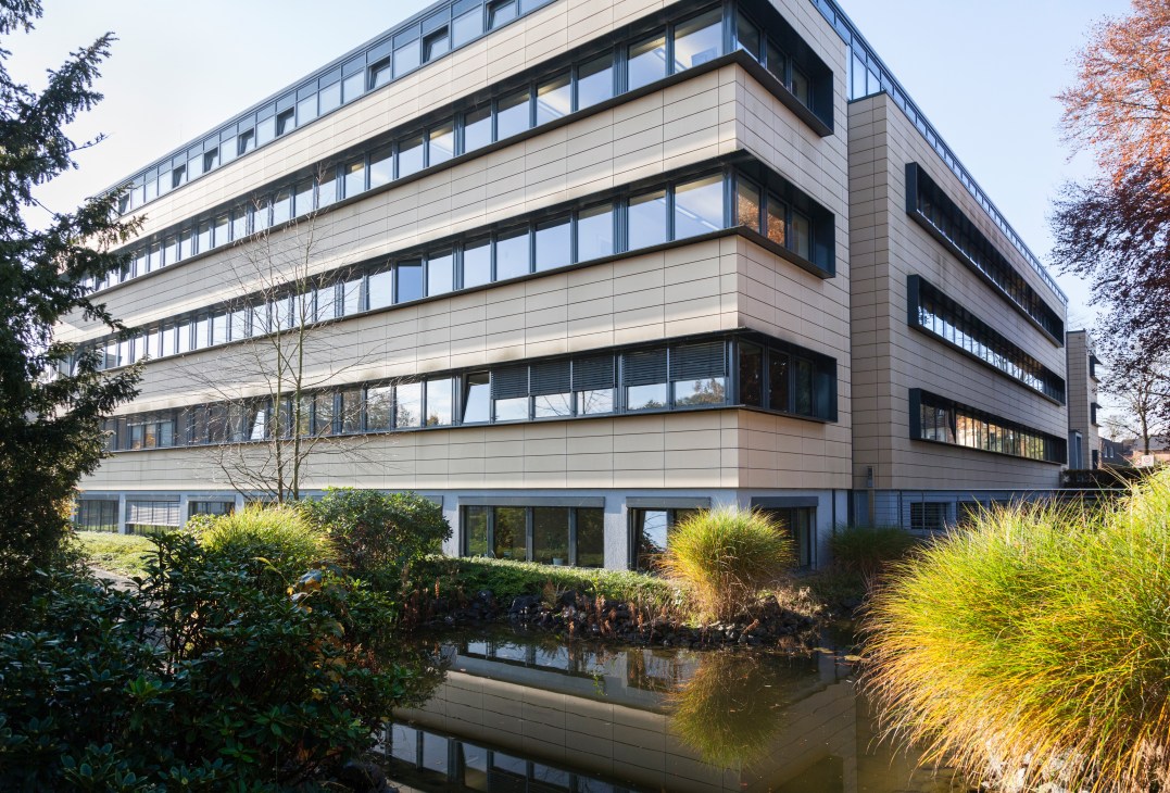 Modern five-storey office building with beige cladding and horizontal bands of windows, surrounded by landscaping with ornamental grasses and shrubs.