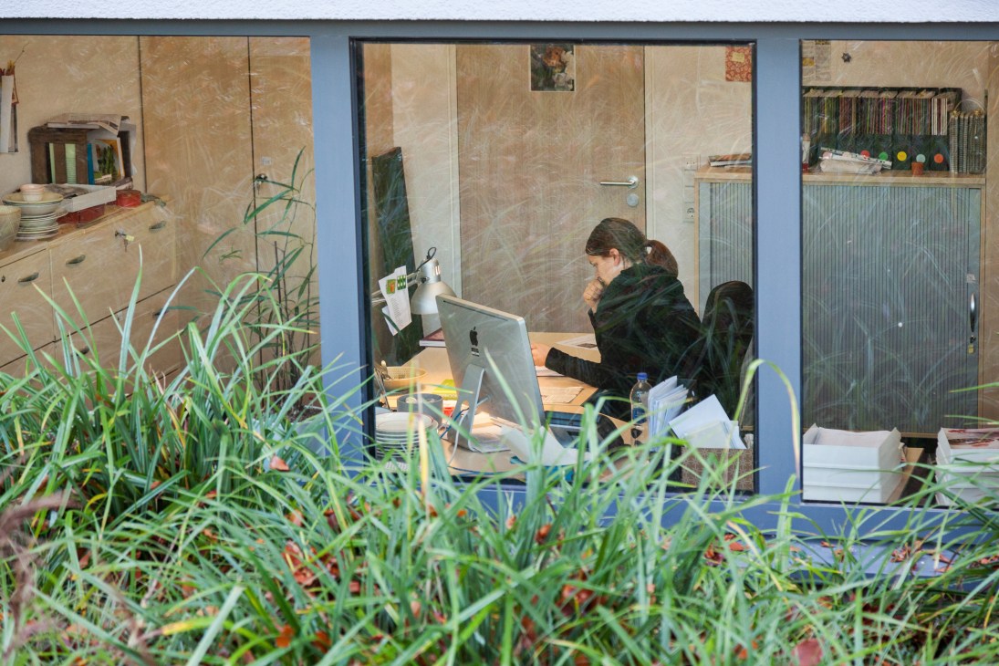 Woman working at desk with computer in Landlust magazine office with plants in foreground.