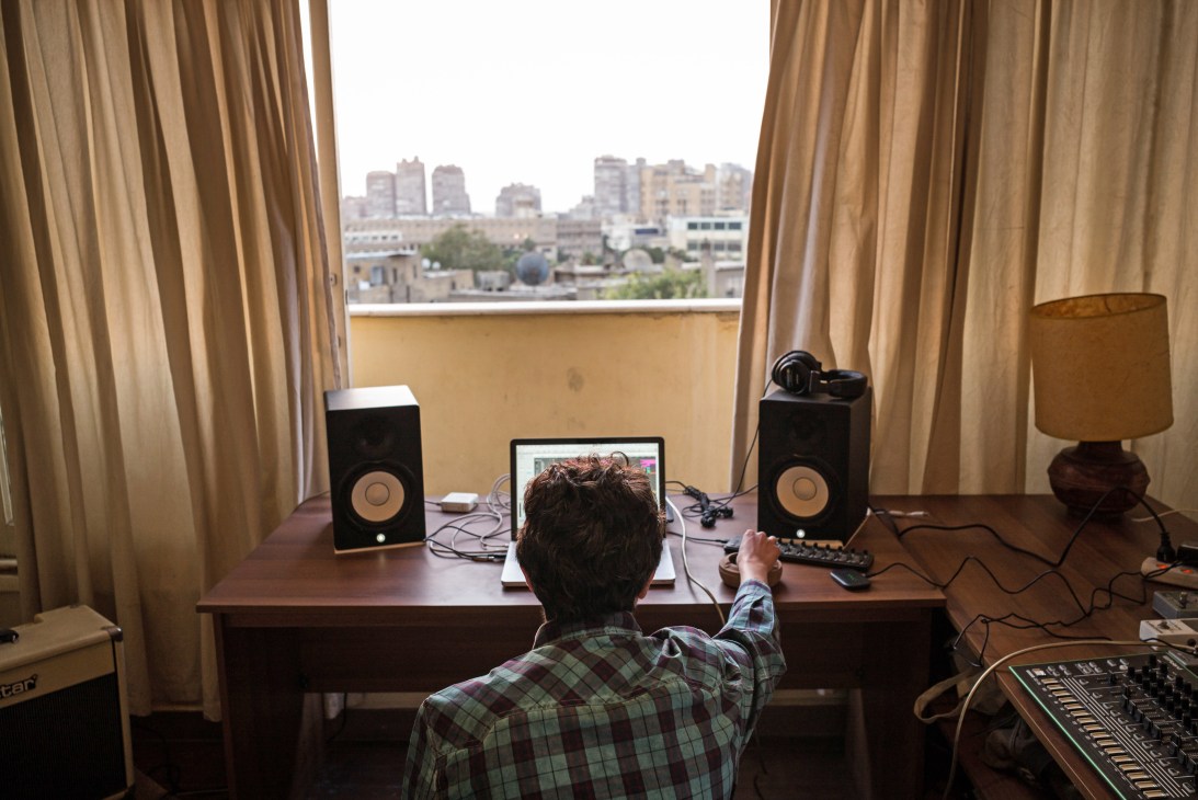 Abdel-Rahman Hussein sits at a desk with a laptop and studio monitors, overlooking a Cairo cityscape through floor-to-ceiling windows.