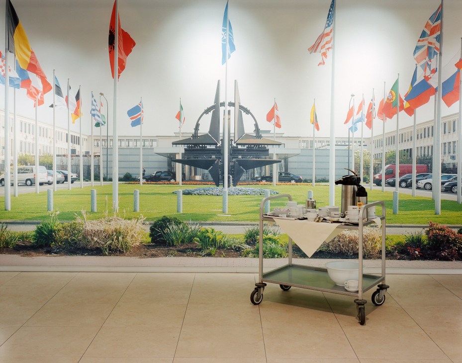 A tea cart with a thermos and cups sits in a bright corridor overlooking NATO flags and headquarters grounds.