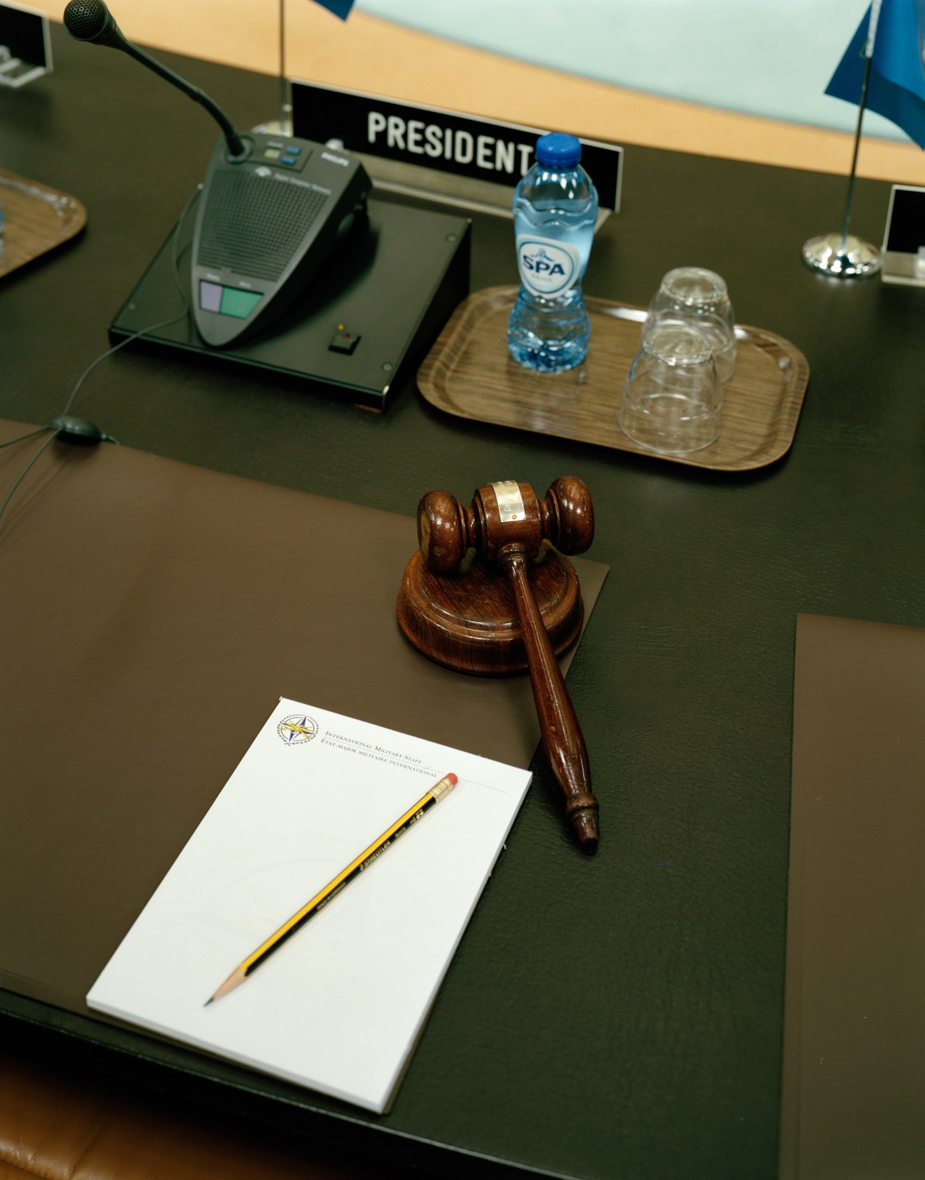 A wooden gavel on a dark green conference table with a microphone, water bottle, and notepad in the North Atlantic Council room.
