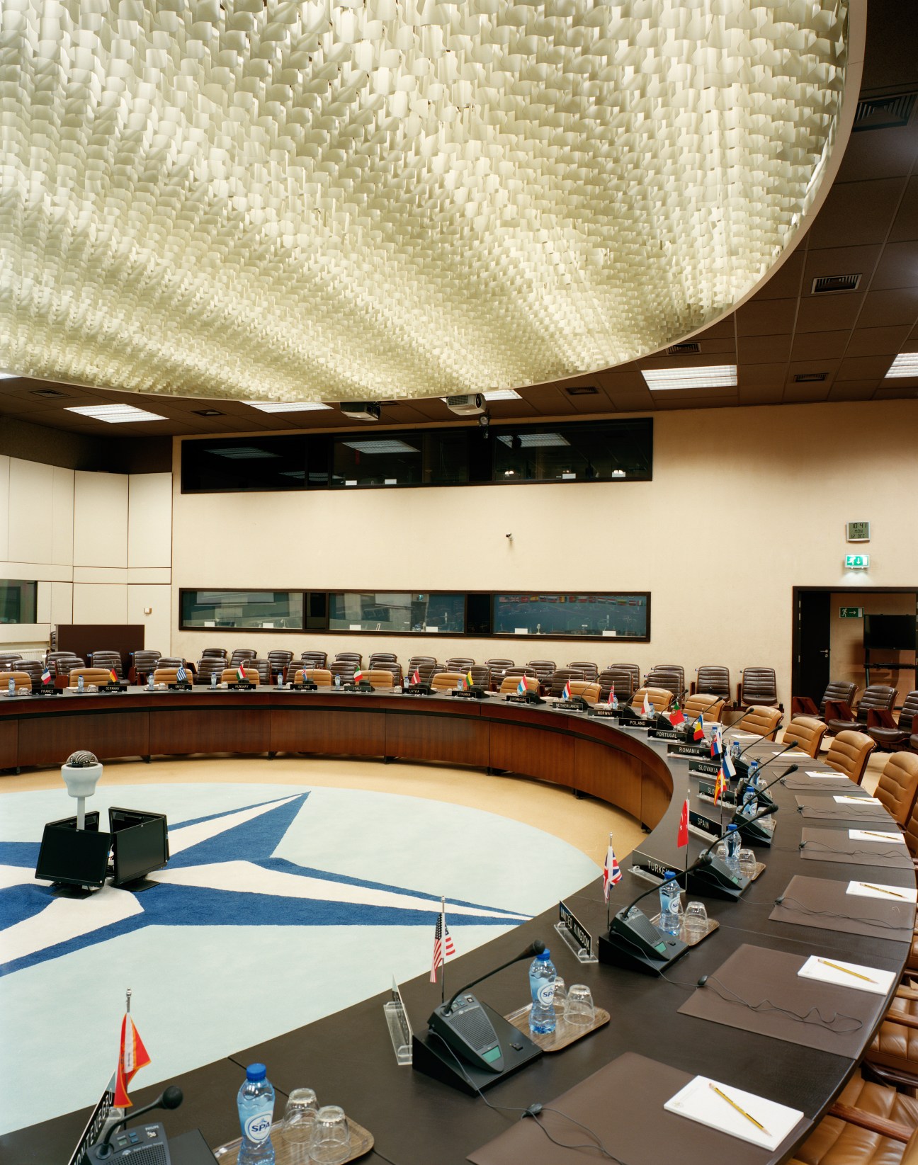 NATO North Atlantic Council chamber with curved seating, country flags, and distinctive woven ceiling.