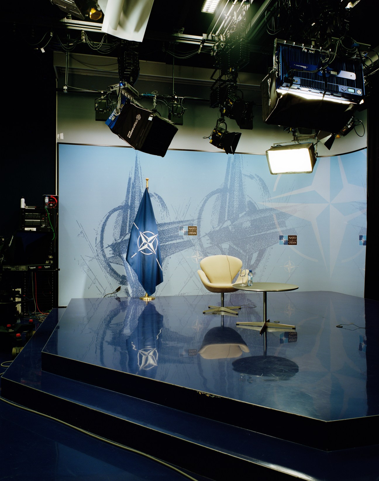 Professional television studio with NATO flag, cream chair, and broadcast lighting equipment for remote interviews.