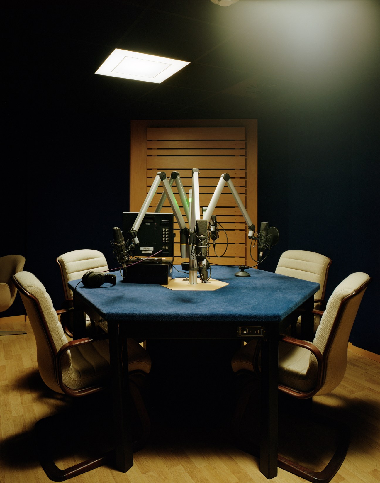 A radio broadcast studio with microphones, cream chairs, and blue hexagonal table in a dark room.