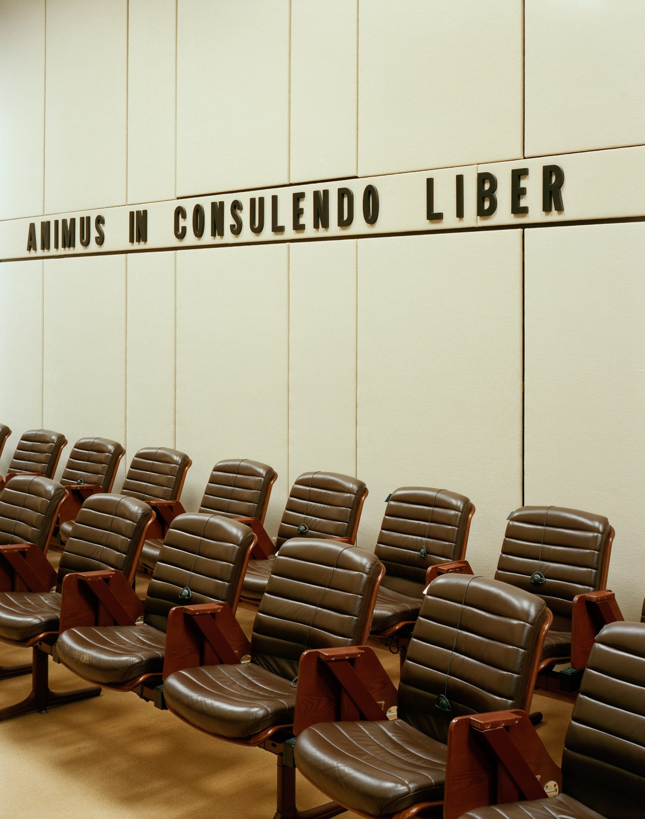 Rows of brown leather chairs in a conference room with Latin motto on cream-coloured wall.