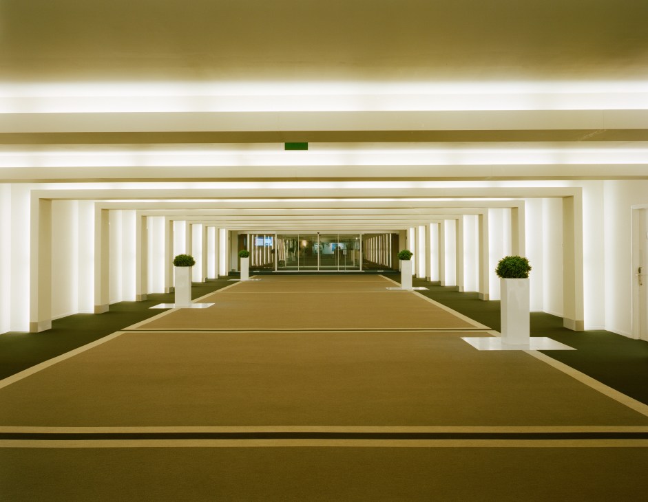 Long modern corridor with white columns, green carpet, and potted plants leading to glass doors in Nato headquarters.