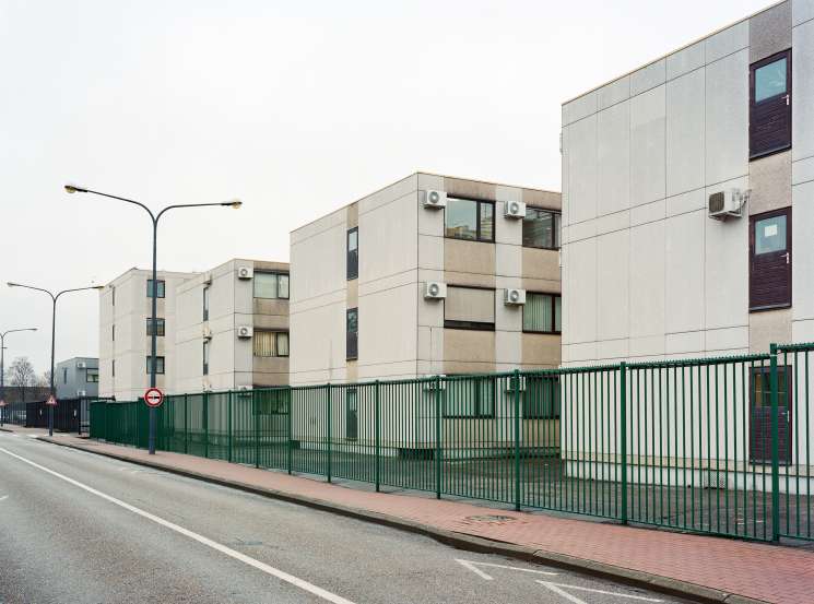 Stacked concrete prefabricated units with air conditioning units and windows, surrounded by a green metal fence.