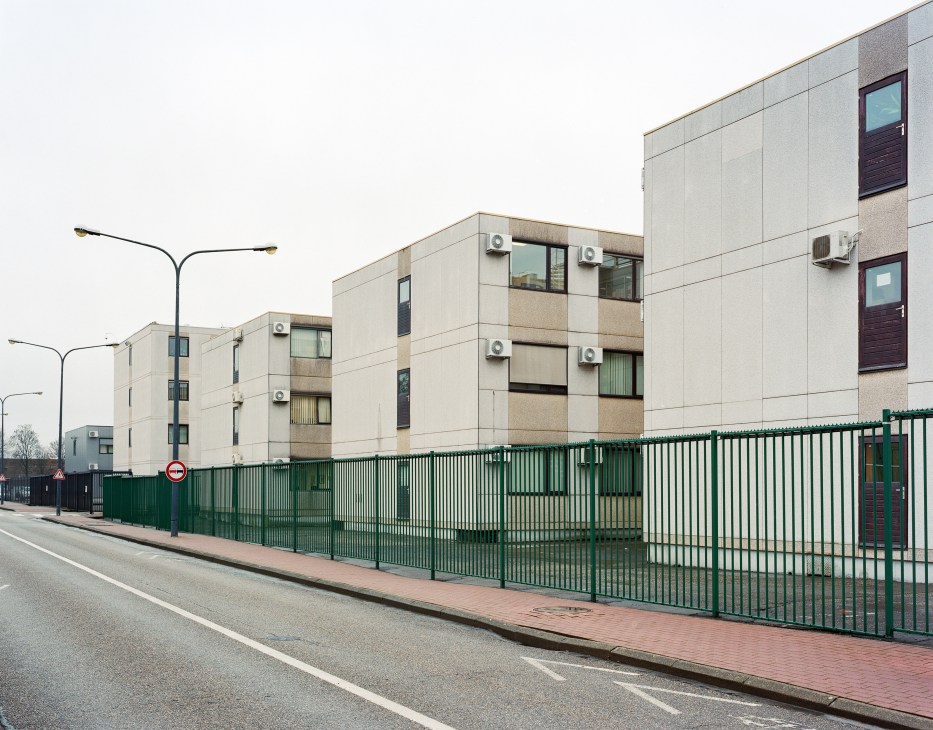 Stacked concrete prefabricated units with air conditioning units and windows, surrounded by a green metal fence.