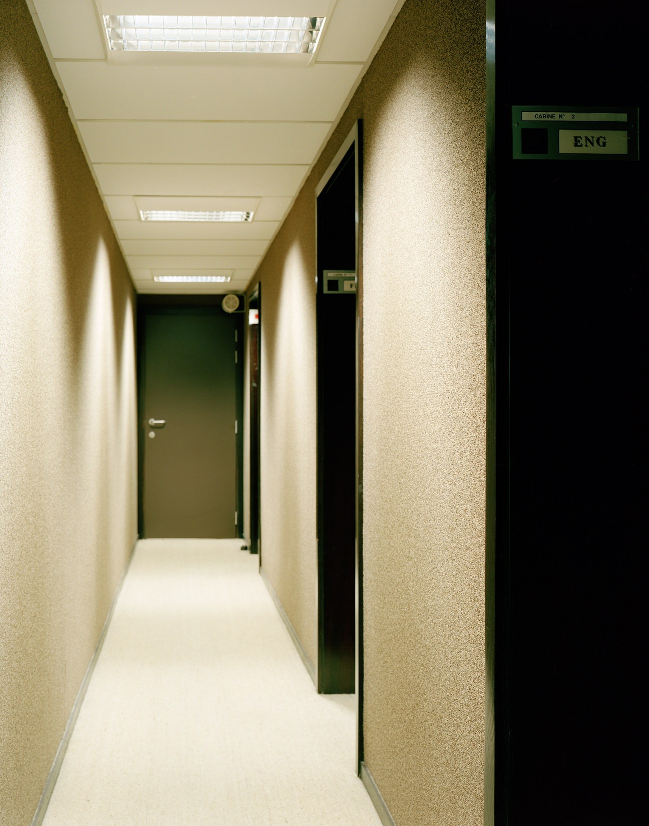 A narrow corridor with beige carpet-covered walls, fluorescent ceiling lights, and a green door marked 'CABINE N° 2 ENG'.