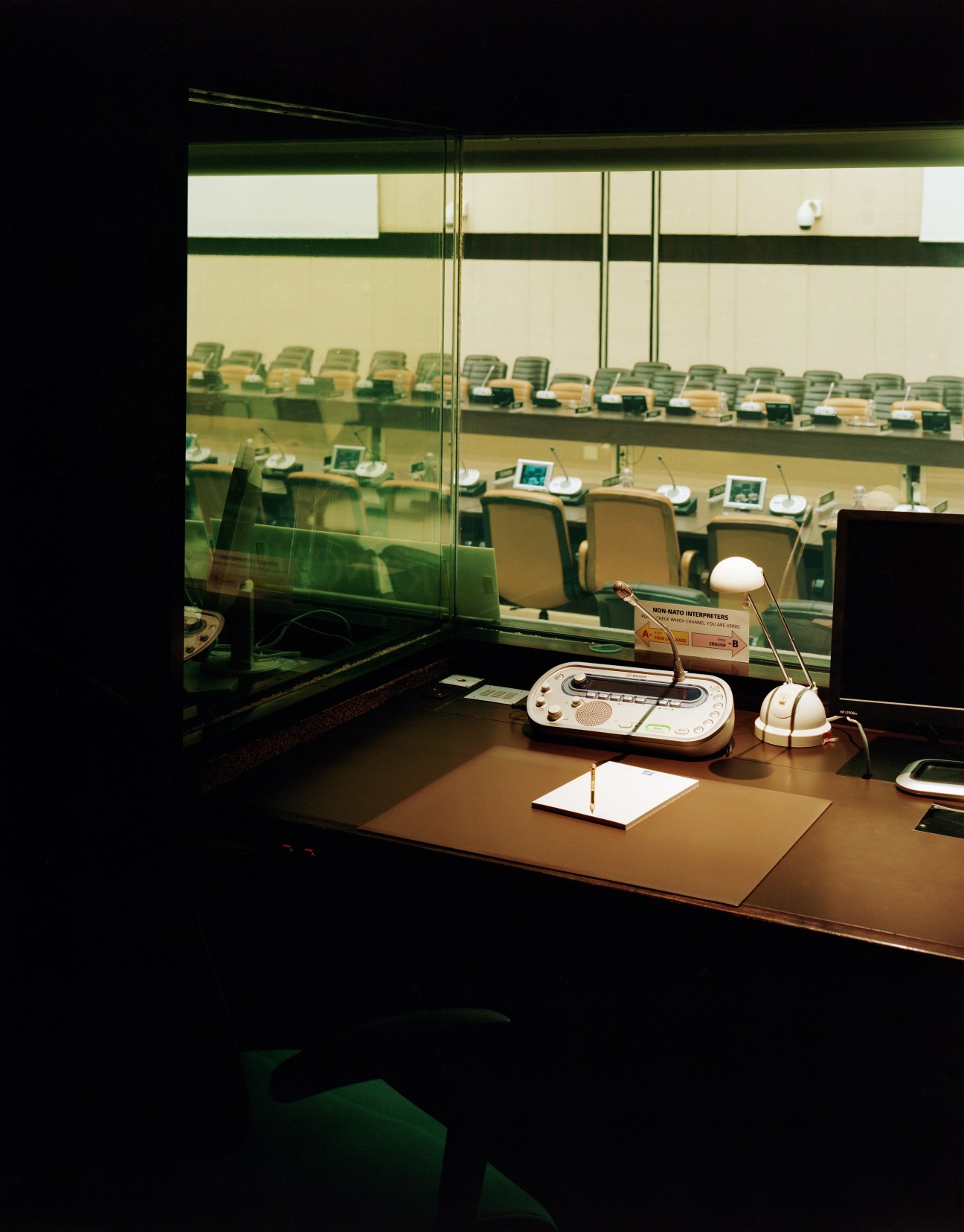 View from a translator's booth overlooking the North Atlantic Council room with rows of green chairs and interpretation equipment.