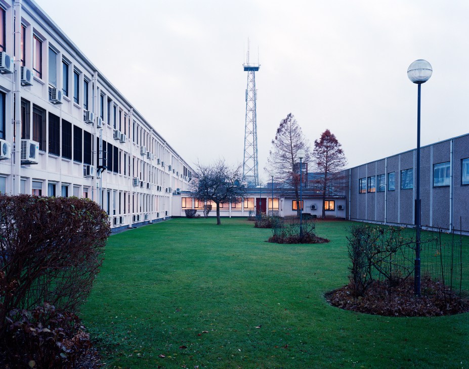 NATO headquarters courtyard with green lawn, brutalist concrete buildings, communication tower and bare trees at dusk.