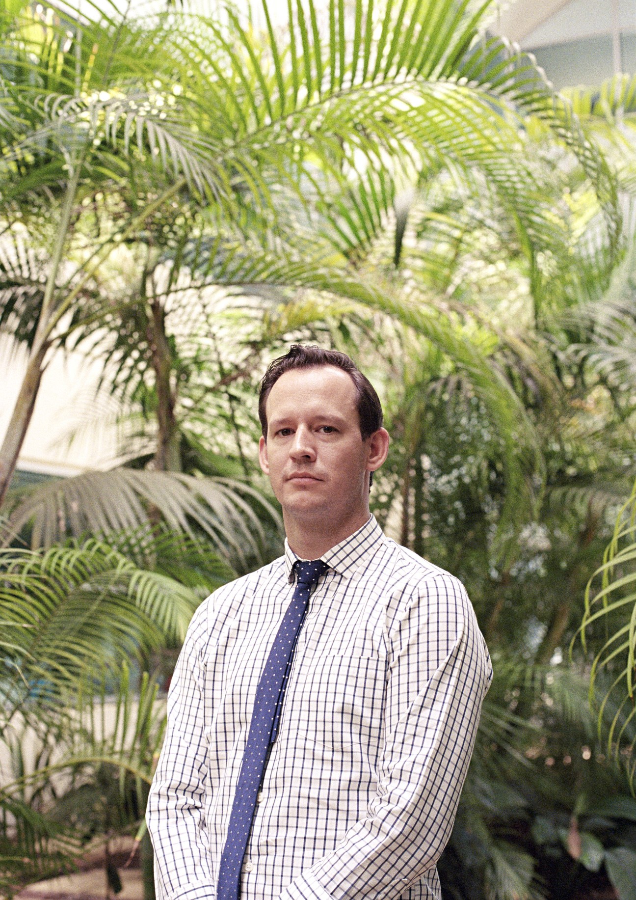 State political editor Gareth Parker standing in front of palm plants wearing a checkered shirt and navy tie.