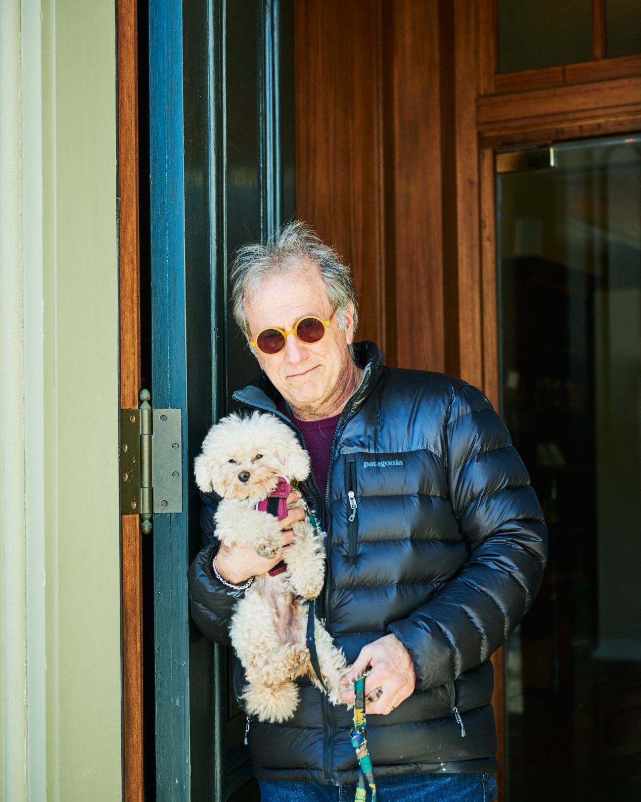 Man with grey hair and round sunglasses holds a small white dog while standing in a doorway.