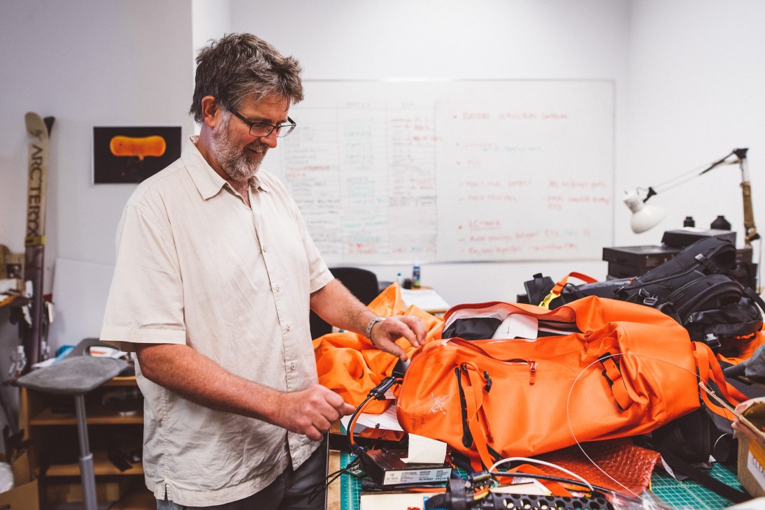 Designer examines bright orange Arc'teryx outdoor gear in a studio workspace with whiteboards and design materials.