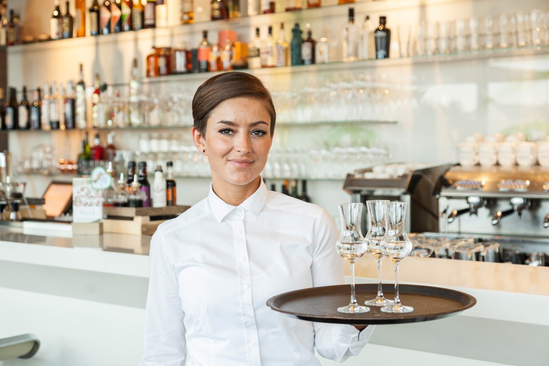 Bartender at Faces aperitif bar holding tray with champagne glasses, shelves of bottles in background.