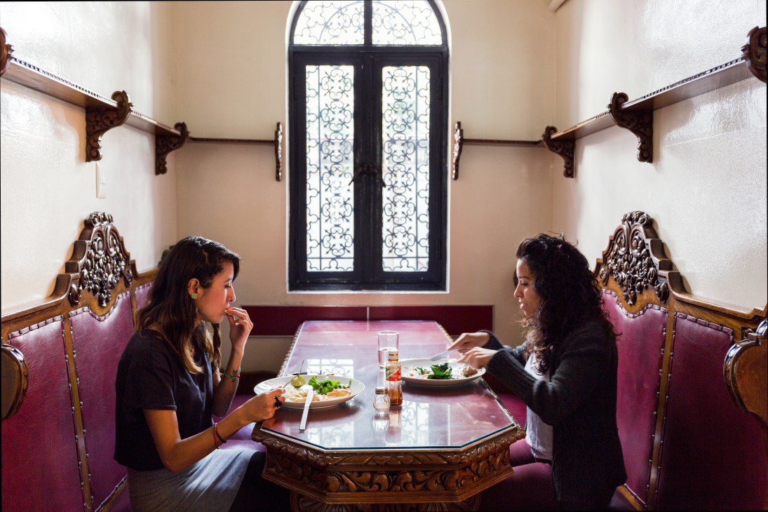 Two staff members having lunch at a table in an ornately decorated office dining area with pink velvet chairs and carved wooden details.